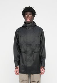 Veste de pluie noire avec capuche, deux poches avant, taille à cordon de serrage et finition lisse et imperméable. Conçue pour un ajustement décontracté.