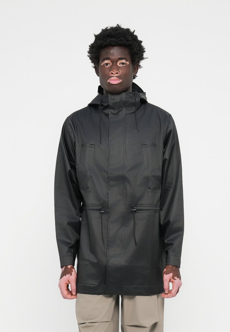 Veste de pluie noire avec capuche, deux poches avant, taille à cordon de serrage et finition lisse et imperméable. Conçue pour un ajustement décontracté.