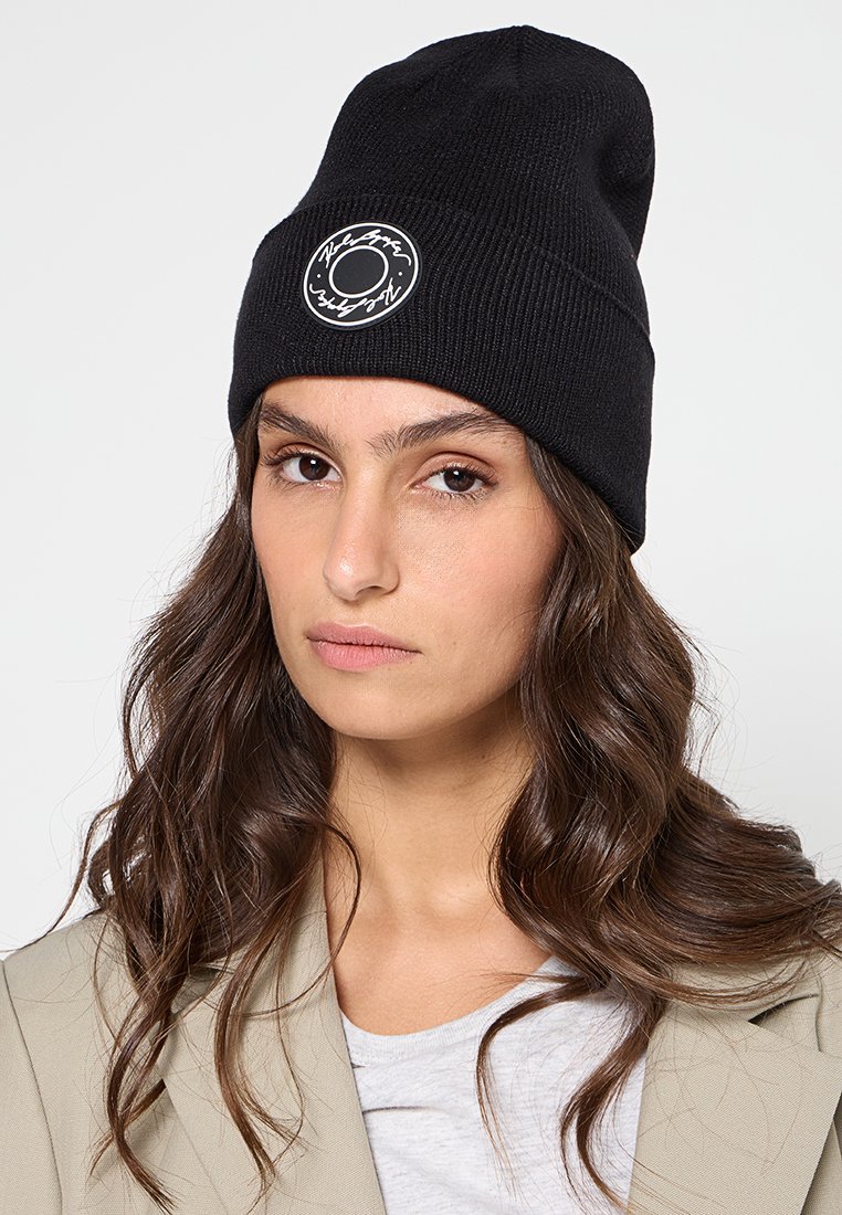 Czarna dzianinowa czapka beanie z prążkowaną teksturą i okrągłą łatą z logo, na której znajduje się biały tekst. Czapka ma luźny krój.