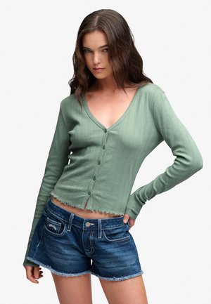 Jeune femme portant un cardigan côtelé vert boutonné et un short en jean bleu foncé aux bords effilochés, posant avec une main sur la hanche.