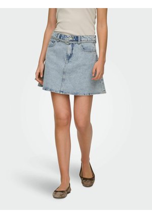 Žena kráča v svetlomodrej denimovej mini sukni s opaskom, béžovom topánku a hnedých balerínach na jednoliatom pozadí.