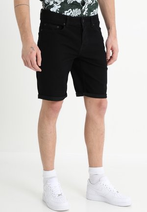 Man draagt zwarte shorts met opgerolde zoom, witte sneakers, witte sokken en een donker bloemenhemd, staand tegen een effen lichte achtergrond.