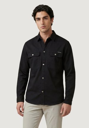 Mand iført sort skjorte med knapper og klaplommer samt lyse beige bukser, stående foran en ensfarvet baggrund