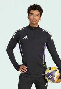 Schwarzer athletischer Pullover mit Viertel-Reißverschluss, weißem Adidas-Logo, lila und grünen Akzenten, langen Ärmeln und einem Fußball in der Hand.