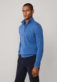 Suéter azul de punto con cuello de media cremallera y puños acanalados. Presenta un logo sutil en el pecho. Lleva puesto sobre pantalones oscuros.