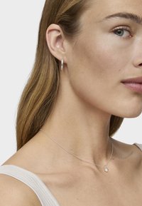 Mujer joven con cabello castaño claro que lleva un pequeño pendiente de aro de plata y un delicado collar de plata con un colgante redondo, vistiendo una blusa sin mangas blanca.