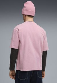 Uomo di spalle che indossa un berretto rosa, una maglietta a maniche corte di un rosa chiaro sopra una maglietta a maniche lunghe nera e jeans blu scuri, su uno sfondo grigio.