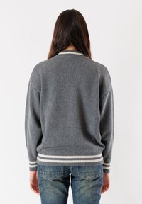 Persona con lunghi capelli scuri che indossa un maglione grigio con strisce bianche sui polsini e sull'orlo, abbinato a jeans in denim blu, girata di spalle.