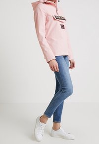 Jasnoróżowa kurtka wiatrówka z przednią kieszenią, czarnym nadrukiem oraz naszywką z flagą Norwegii. Noszona z niebieskimi dżinsami i białymi trampkami.