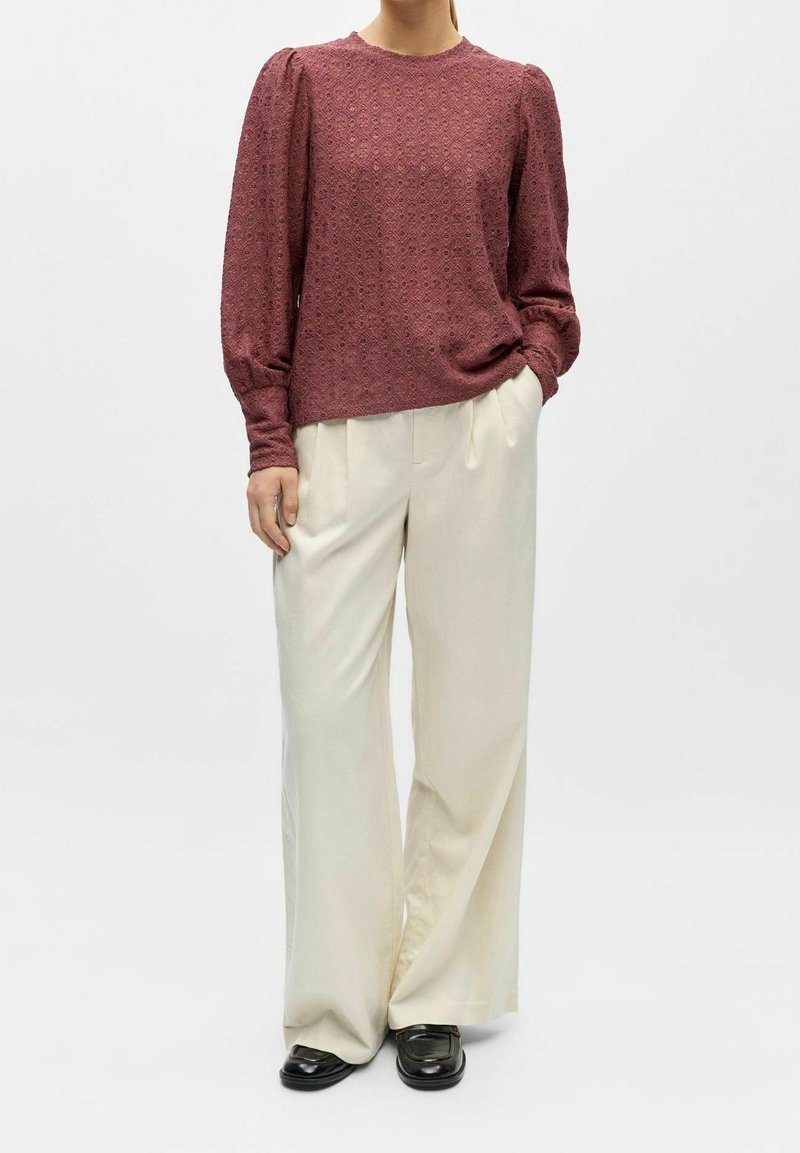 Personne portant un chemisier bordeaux texturé à manches longues, pantalon crème à jambes larges, mocassins noirs, debout avec une main dans la poche sur fond blanc.