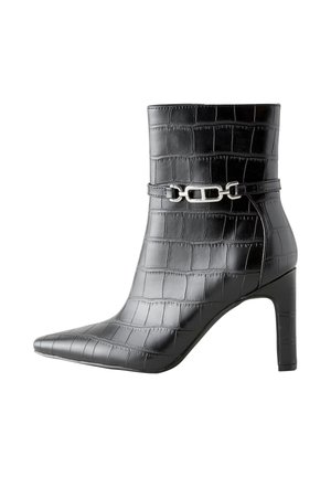 Beine in einem schwarzen Rock, die glänzende schwarze spitze Stiefeletten mit einem Krokodilmuster tragen, stehen auf einem grauen Boden.