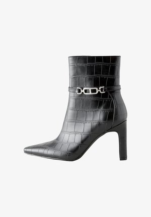 Piernas con una falda negra que llevan botas negras brillantes de punta puntiaguda con un patrón de cocodrilo, de pie sobre un suelo gris.