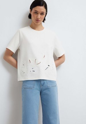 Donna con capelli corti e scuri indossa una maglietta bianca a maniche corte con intagli floreali e jeans azzurri a vita alta, in piedi contro un muro bianco.