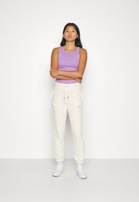 Top senza maniche viola abbinato a pantaloni joggers crema. I joggers presentano un elastico in vita e polsini alla caviglia. Il modello indossa sneaker bianche.