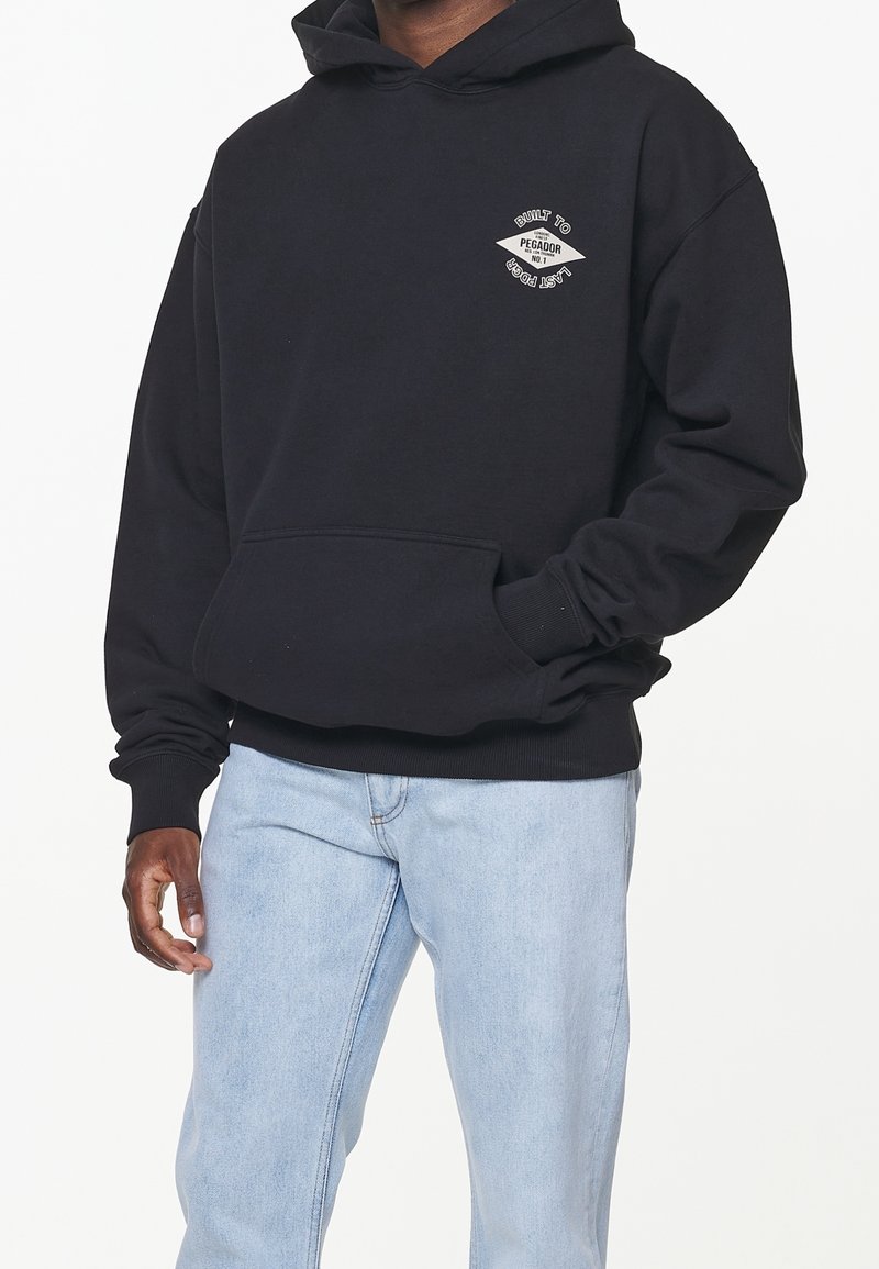 Hombre con sudadera negra con bolsillo frontal y pequeño logo blanco en el pecho, combinado con jeans azul claro, de pie ante un fondo blanco.