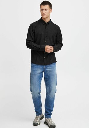 Black button-up shirt made of fabric with a fine texture, paired with light blue denim jeans and grey athletic shoes, standing on a plain background.