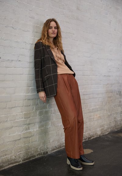 Blazer xadrez preto sobre uma blusa bege, combinado com calças na cor ferrugem e sapatos de plataforma pretos, em frente a uma parede de tijolos de cor clara.