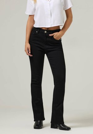 Kobieta ubrana w czarne jeansy bootcut z przednimi kieszeniami, białą krótką koszulę zapinaną na guziki oraz czarne botki na obcasie, stojąca na neutralnym tle.
