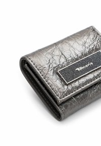 Cartera plateada de cuero sintético arrugado con un adorno rectangular negro que presenta el nombre de la marca "Tamaris" en letras plateadas.