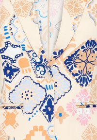 Blazer beige avec des motifs floraux et géométriques bleus, pêches et roses, doté d'un bouton blanc unique et de rabats de poche.