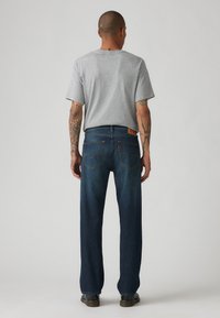 Grijze T-shirt met korte mouwen, gecombineerd met donkerblauwe straight-leg jeans, voorzien van een leren patch en zichtbare stiksels op de achterzakken.