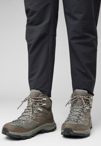 Stivali da trekking marroni con tessuto testurizzato, occhielli metallici e lacci riflettenti. La suola in gomma offre trazione. Abbinati a pantaloni scuri e a sigaretta.