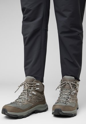 Braune Wanderschuhe aus strukturiertem Stoff, mit Metallösen und reflektierenden Schnürsenkeln. Gummisohlen sorgen für Traktion. Kombiniert mit dunklen, schmalen Hosen.