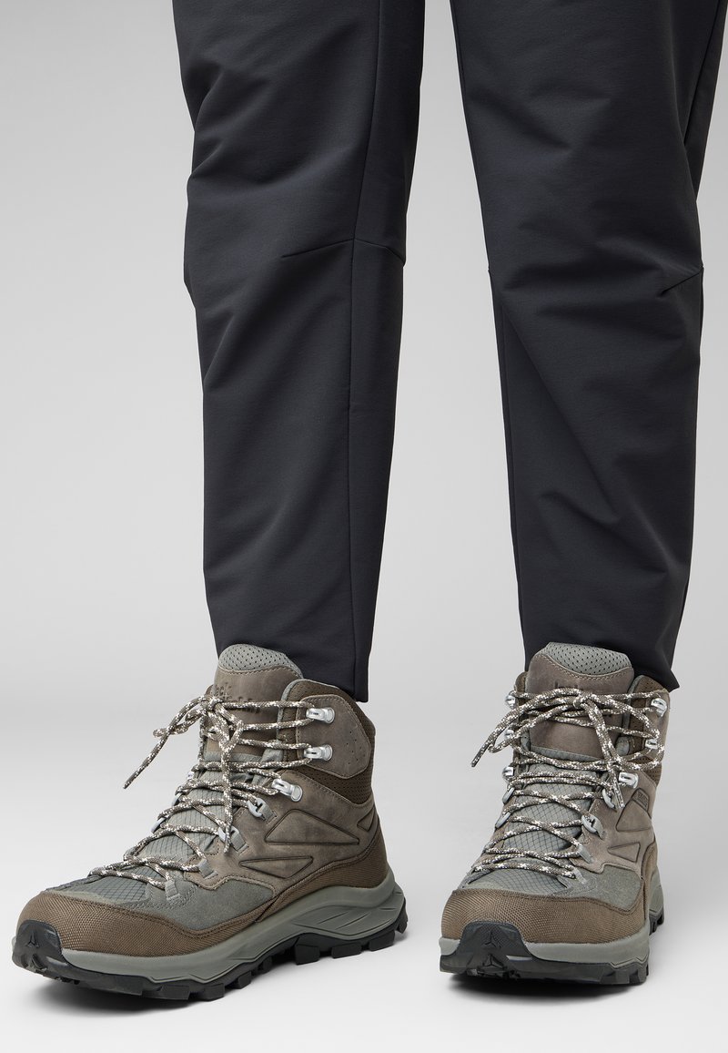 Stivali da trekking marroni con tessuto testurizzato, occhielli metallici e lacci riflettenti. La suola in gomma offre trazione. Abbinati a pantaloni scuri e a sigaretta.