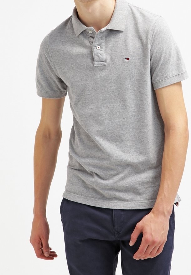 Homme portant un polo gris clair à manches courtes et un pantalon bleu foncé, se tenant devant un fond blanc uni.