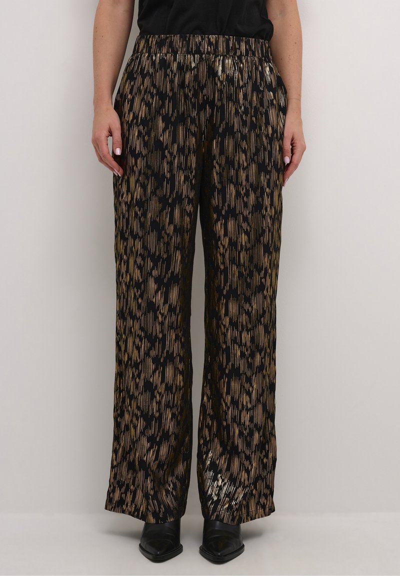 Femme portant un pantalon large à motifs noirs et dorés, des chaussures noires à bout pointu et un haut noir, sur un fond uni.
