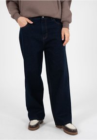 Vaqueros de mezclilla en azul marino oscuro con un corte holgado, que presentan un diseño tradicional de cinco bolsillos y costuras naranjas contrastantes.