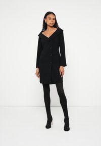 Melna mētelis ar platu apkakli, piegulošu formu un priekšējiem pogām. Aprīkots ar garām piedurknēm un sānu kabatām papildus funkcionalitātei.