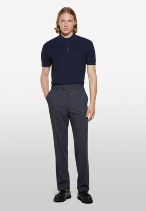 Polo shirt navy à surface texturée, à manches courtes, avec un plastron à trois boutons, associé à un pantalon gris ajusté et des chaussures noires.