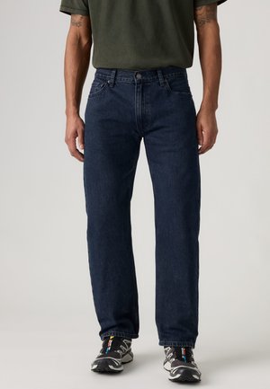 Homme portant un jean droit bleu foncé, des baskets noires et blanches, et un t-shirt vert olive, debout devant un fond uni.