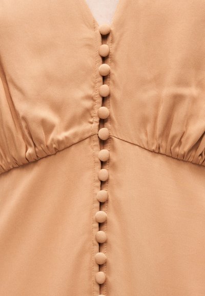 Κοντινό πλάνο μπεζ υφασμάτινου φορέματος με πτυχωτή λεπτομέρεια στο στήθος και μια κάθετη σειρά από καλυμμένα στρογγυλά κουμπιά στο κέντρο.