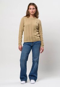 Jeune femme debout portant un pull beige en maille torsadée, un jean évasé bleu et des baskets blanches sur un fond clair uni.