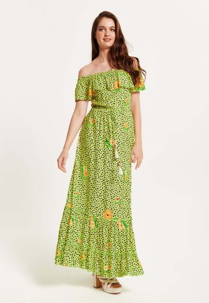 Green off-shoulder maxi dress with black polka dots and floral patterns. Features a belted waist and a ruffled hem. Smooth fabric and flowy design.