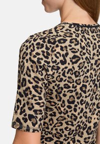 Top en tissu côtelé à motif léopard, avec des manches courtes et un col arrondi, sur un fond beige agrémenté de taches noires et marron.