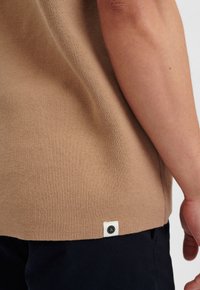 Beiger Pullover aus weichem Strickstoff, mit einem Etikett am Saum und einem schwarzen Logo, kombiniert mit dunkel gefärbten Hosen.