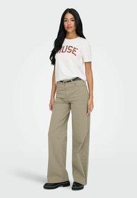 Femme aux longs cheveux noirs portant un T-shirt blanc "MUSE", un pantalon beige à jambes larges, une ceinture noire et des chaussures noires épaisses sur fond clair.