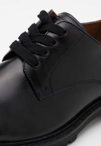 Chaussure en cuir noir avec bout rond, présentant un laçage noir, des coutures discrètes et une semelle en caoutchouc texturé pour une adhérence et une durabilité accrues.