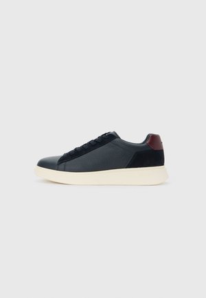 Black low-top lace-up sneaker with textured leather front, suede heel, burgundy heel tab, and thick off-white sole against light background.