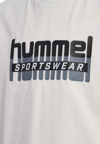 Witte katoenen t-shirt met een opvallende zwart-blauwe grafiek met de tekst "hummel" en "SPORTSWEAR", en een gestructureerd stippenpatroon op de achtergrond.