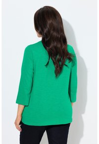 Top verde con maniche a 3/4 in tessuto leggermente strutturato. Sul retro presenta una scollatura arrotondata e una vestibilità morbida. Indossato con denim scuro.