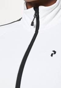 Witte zip-up jas met hoge kraag, voorzien van een zwarte rits en logo. Glad materiaal met minimale textuur en zwarte accenten.