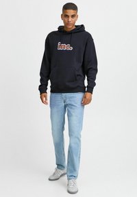 Sudadera azul marino con bolsillo frontal, presenta el logotipo "inc." en letras de colores, combinada con jeans azul claro y zapatillas grises.
