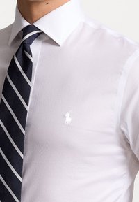 Biała koszula z klasycznym kołnierzykiem, z małym białym haftowanym logo, w zestawie z granatowym krawatem w paski. Gładka tekstura materiału.