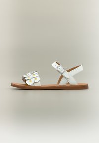 Sandale blanche plate avec semelle marron, boucle de sangle cheville, et bride avant décorée de fleurs blanches superposées avec centres jaunes.