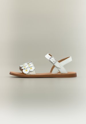 Bílá plochá sandála s hnědou podrážkou, přezkou na kotníkovém pásku a předním páskem ozdobeným vrstvenými bílými květy se žlutými středy.
