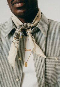 Detailaufnahme einer Person, die einen beigefarbenen, gemusterten Schal um den Hals trägt, ein gestreiftes Button-Up-Hemd und zwei goldene Halsketten.